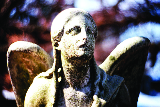 A photograph is shown of an angel statue. While some details of the statue, including facial features, are present, effects of weathering appear to be diminishing these features.